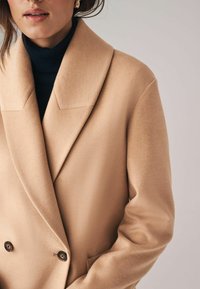 Beige Doppelreiher mit weicher Textur, breitem Reverskragen und dunklen Knöpfen. Trägt man über einem schwarzen Rollkragenpulli.
