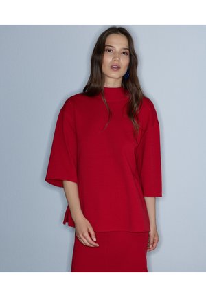 Mujer con cabello largo y castaño que lleva una blusa roja suelta con mangas hasta el codo y una falda roja a juego, de pie frente a una pared gris clara.