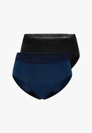 Femme portant un crop top noir et un slip taille haute bleu marine, posant avec un bras levé derrière la tête sur un fond blanc.
