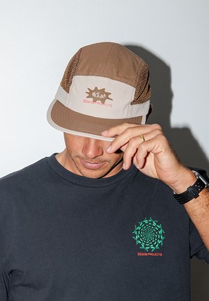 Άνδρας που φορά καφέ και μπεζ καπέλο με λογότυπο, μαύρο μπλουζάκι με πράσινο σπειροειδή σχεδιασμό και κόκκινο κείμενο, ρυθμίζει το γείσο του καπέλου με το δεξί του χέρι.