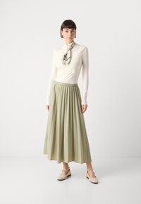 Lys beige langermet topp, grønn plisse midi-skjørt og bleke flate sko. Skjerf med blomsterdesign bundet rundt halsen, jevn tekstur generelt.