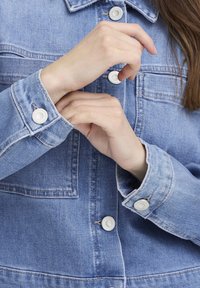 Világoskék farmerdzseki ezüst patentokkal, látható varrással és laza fazonnal; az ujjvégek finoman fel vannak hajtva, így látható a belső anyag textúrája.