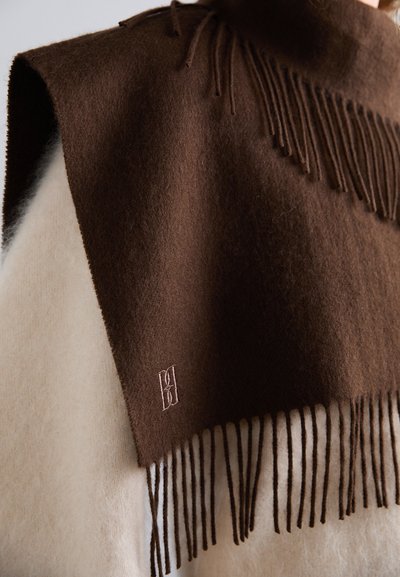 Écharpe en laine marron avec des bords frangés, présentant un léger motif texturé. Un logo brodé est visible près du bord. Portée sur un vêtement crème clair.