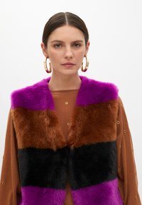 Faux fur vest with horizontal color blocks: purple, brown, black, and purple. Soft texture with a wide collar. Worn over a brown top.