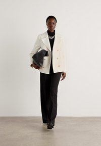 Dubbelgeknoopt witte blazer met gouden knopen, zwarte coltrui, brede zwarte broek en een zwarte slouchy tas, gezet tegen een neutrale achtergrond.