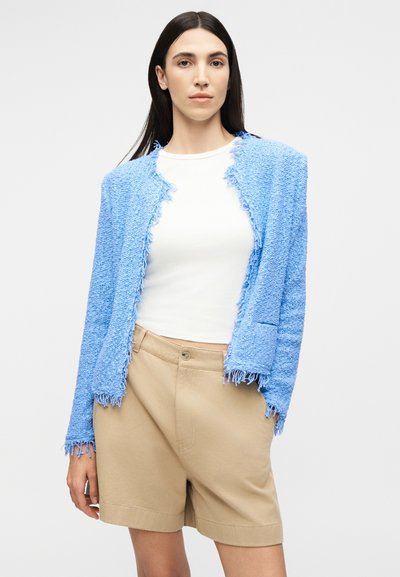 Γυναίκα που φοράει ανοιχτό μπλε σακάκι με υφή πάνω από λευκό τοπ και μπεζ σορτς ψηλόμεσο, στέκεται μπροστά σε απλή φόντο.