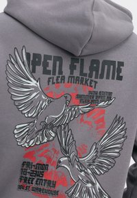 Szara bluza z kapturem z dużym nadrukiem przedstawiającym dwa stylizowane ptaki, napis "OPEN FLAME FLEA MARKET" oraz czerwone plamy akcentujące.
