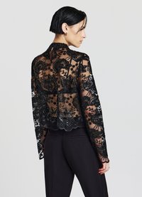 Giacca nera in pizzo con ricami floreali e orlo ondulato, con maniche lunghe e collo alto, indossata sopra pantaloni neri.