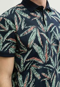 Marineblauwe polo met een textuur van botanisch patroon in groen en koraal. Voorzien van een donkere kraag en knopen, die het gedurfde ontwerp accentueren.