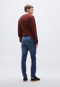 Férfi bordázott burgundi pulóver kék farmernadrággal párosítva. Kiemelkedő részletek közé tartozik a barna bőrből készült márkajelzés a derékpánton és a fekete cipők.