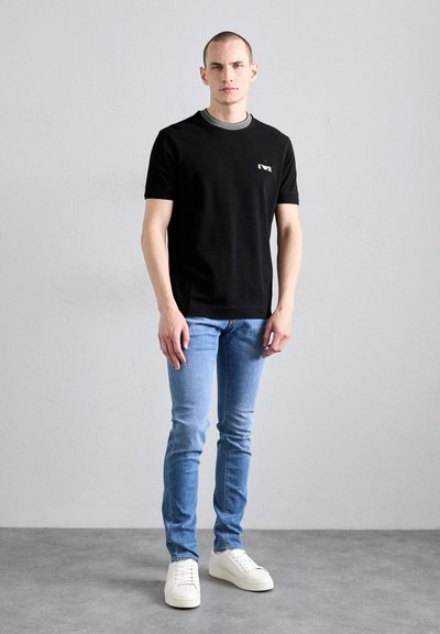 T-shirt noir avec un col rond et un petit logo blanc sur la poitrine, associé à un jean léger bleu ajusté et des baskets blanches.