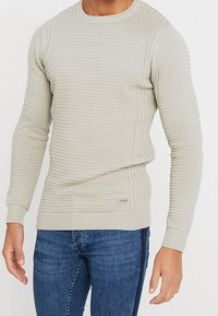 Ljusbeige stickad tröja med ribbad textur, rund halsringning och åtsittande ärmar. Har en sidolapp nära fållen. Kombineras med blå jeans.