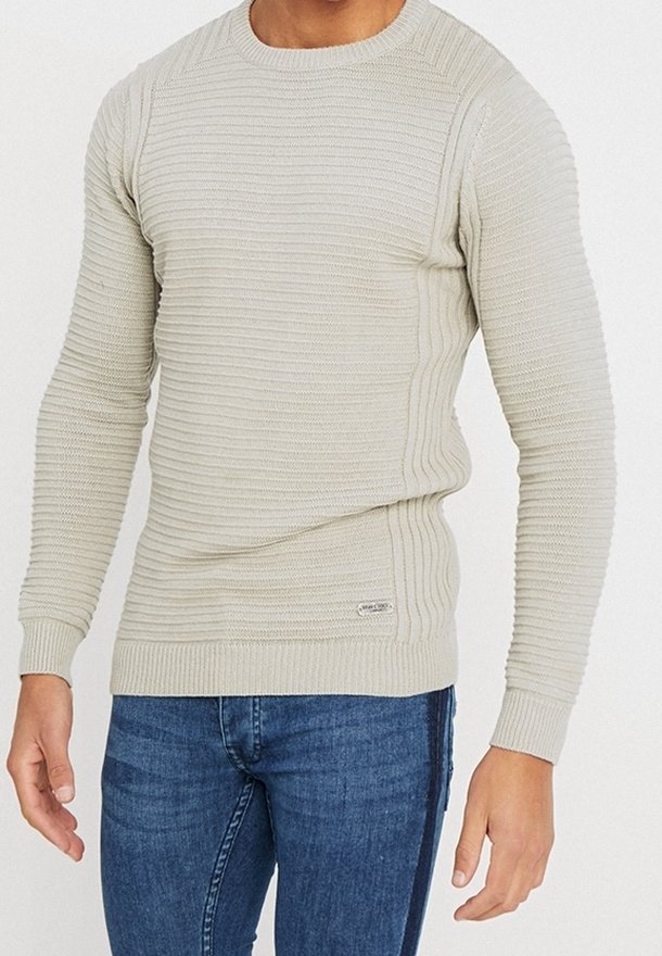 Ljusbeige stickad tröja med ribbad textur, rund halsringning och åtsittande ärmar. Har en sidolapp nära fållen. Kombineras med blå jeans.
