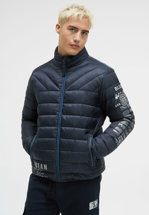 Gepolsterte marineblaue Jacke mit hohem Kragen, Seitentaschen, blauen Reißverschluss und weißer Markenaufschrift entlang der Ärmel. Glatte, glänzende Textur.