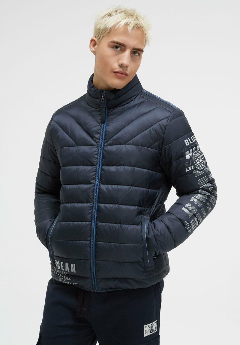 Gepolsterte marineblaue Jacke mit hohem Kragen, Seitentaschen, blauen Reißverschluss und weißer Markenaufschrift entlang der Ärmel. Glatte, glänzende Textur.