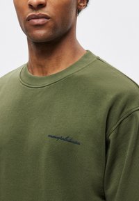 Sudadera verde oliva con cuello redondo, que presenta un cuello acanalado y texto bordado en azul marino en el frente. Tejido suave y texturizado.