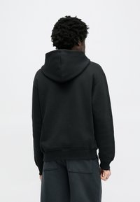 Μαύρο φούτερ με κουκούλα, μαλακή υφή, ριμπ μανσέτες και ζώνη μέσης, φορεμένο με σκούρο παντελόνι. Οπίσθια όψη σε απλό φόντο.