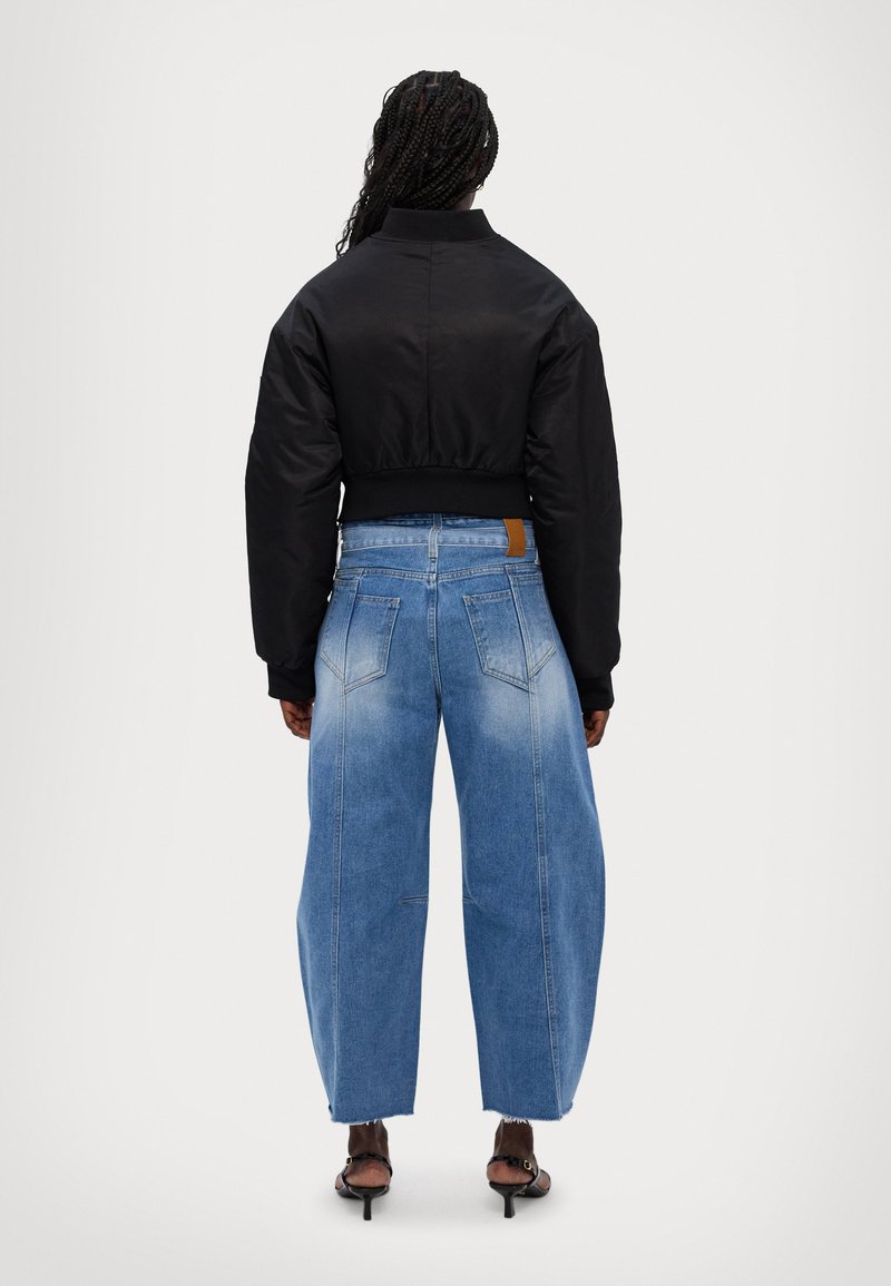 Zwarte cropped bomberjack met brede mouwen, gedragen met hooggepolijste lichtblauwe wijde jeans. Opvallend tan leren label op de jeans.