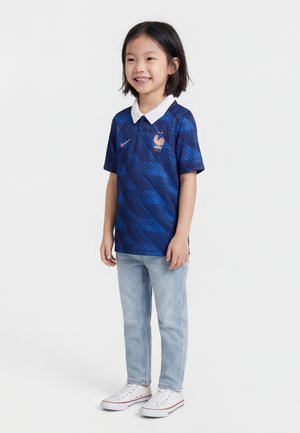 Dziecko stoi i się uśmiecha, ubrany w niebieską koszulkę reprezentacji Francji w piłce nożnej z białym kołnierzykiem, jasnoniebieskie dżinsy i białe trampki na jednolitym tle.