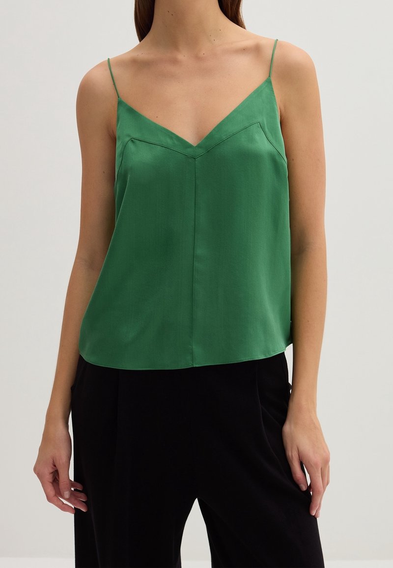 Camisole en satin vert avec des bretelles fines, décolleté en V, coupe ajustée et texture lisse, associée à un pantalon noir à coupe ample.