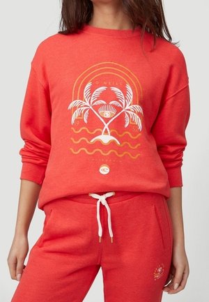 Frau trägt korallenfarbenen Pullover und Hose mit Palmen- und Wellendesign auf dem Pullover und weißen Kordeln an der Hose.