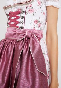 Robe dirndl blanche florale avec corsage burgundy à lacets, crochets argentés décoratifs et nœud en tissu rose brillant attaché à la taille.