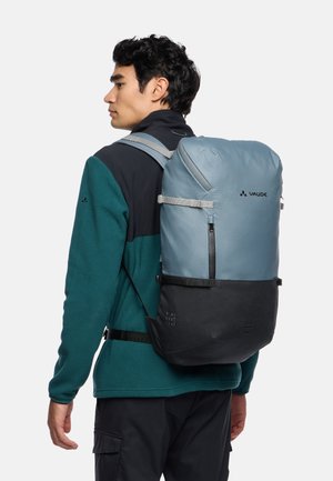 Sac à dos bleu clair avec une texture mate, des sangles grises et un fond noir. Il présente une poche frontale zippée et est marqué "VAUDE."