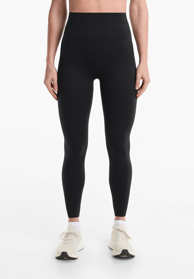 Leggings neri a vita alta realizzati in un materiale liscio e elastico. Presentano un design senza cuciture per una silhouette snella. Abbinati a sneaker leggere.