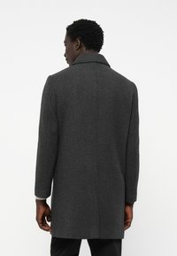 Férfi sötétszürke húsos mintás hosszú kabátban, hátulról látható egy sima fehér háttér előtt.