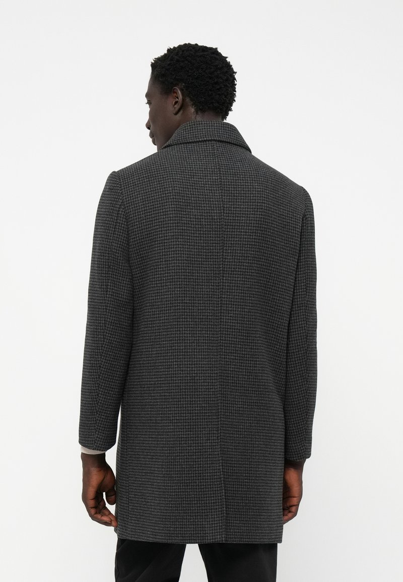 Homme portant un long manteau à motif pied-de-coq gris foncé, vu de dos contre un fond blanc uni.