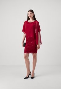 Punainen mekko, jossa on pitsiyksityiskohtia, kerros chiffon-viittaa, aallotettu helma, yhdistetty mustan laukkuun ja korkokenkiin. Yhteispinta on sileä.