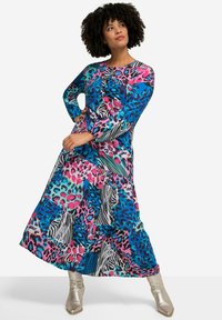 Robe maxi à manches longues arborant un patchwork vibrant de motifs animaliers dans des teintes de bleu, rose et vert, avec un ourlet à volants. Bottines argentées.