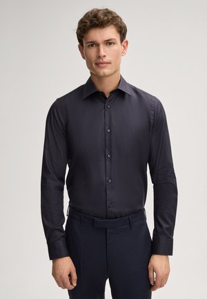 Chemise boutonnière bleu marine foncé à manches longues, avec un motif texturé subtil et des boutons noirs. Coupe ajustée, associée à un pantalon sombre.
