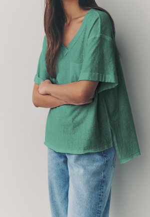 Kobieta z długimi brązowymi włosami, ubrana w luźną zieloną koszulkę z dekoltem w serek i jasnoniebieskie dżinsy, ze skrzyżowanymi ramionami, stojąca na tle białej ściany.