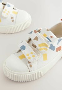 Białe sneakersy z kolorowymi geometrycznymi wzorami, wyposażone w gumowy czubek oraz dwa rzepy do łatwego zapinania. Widoczna teksturowana podeszwa.