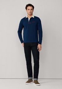 Marineblaues Langarm-Polohemd mit weißem Kragen und rotem Branding. Kombiniert mit dunklen Jeans und braunen Sneakers.