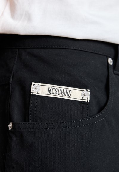 Mustad kangast püksid, mille tagakotil on nähtav valge silt "Moschino", mis on kinnitatud hõbedaste kinnitusdetailide abil ning millel on kontrastne õmblus.