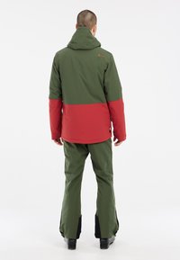 Zimski outfit od dva dijela koji se sastoji od jakne u zelenoj i crvenoj boji s kapuljačom i odgovarajućim zelenim hlačama. Uključuje crne detalje na rubovima hlača.