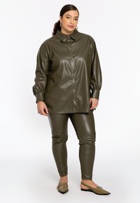 Olijfgroene faux leren blouse met knoopsluiting, oversized silhouet en pofmouwen, gecombineerd met bijpassende strakke broek en puntige flats.