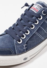 Sininen denimsneaker valkoisella kumipohjalla, metallisilla rei'ittäjillä, tummansinisin nauhoin ja kontrastivärisillä ompeluilla. Merkittävä etiketti kielen päällä.