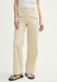 Pantalons larges beiges en tissu léger. Avec une taille à cordon de serrage et des poches avant. Associés à des sandales plates.