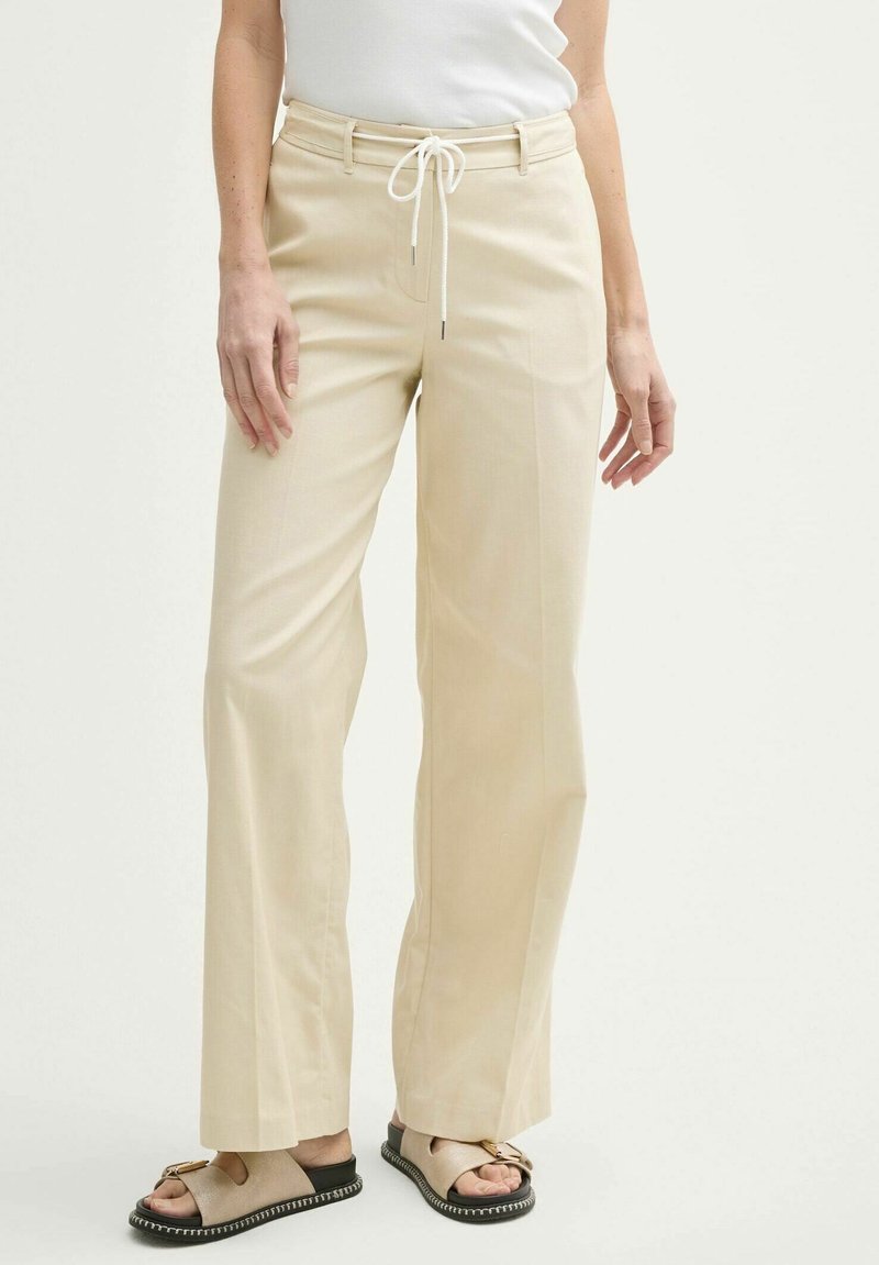 Pantalons larges beiges en tissu léger. Avec une taille à cordon de serrage et des poches avant. Associés à des sandales plates.