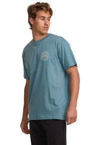 Short-sleeve, crew neck t-shirt in teal cotton with a circular mountain graphic in beige on the left chest. Smooth texture, relaxed fit.