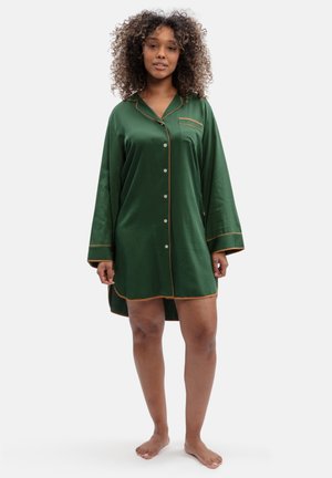 Femme aux cheveux bouclés debout pieds nus portant une longue chemise de nuit boutonnée vert foncé avec un passepoil beige et une poche poitrine.