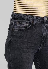Jeans de mezclilla negros con un corte ajustado, diseño de cinco bolsillos, costuras en contraste y cierre de botón. La textura parece ligeramente desgastada.