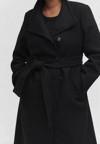 Cappotto in misto lana nero con un colletto prominente, bottoni doppi, cintura e tasche laterali, che trasmette una silhouette strutturata.