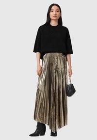 Donna che indossa una blusa nera oversize a maniche corte, una gonna lunga plissettata dorata metallica, stivali neri e tiene in mano una piccola borsa nera con manico a catena dorata.