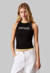 Top aderente nero a costine con spalline gialle, caratterizzato dal logo "CALVIN KLEIN" in bianco. Abbinato a jeans bianchi a vita alta.