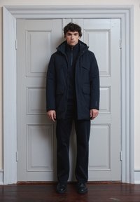 Marine parka met een capuchon, ritssluiting en meerdere voorzakken, gecombineerd met een bijpassende top en broek, voor een lichte deur achtergrond.