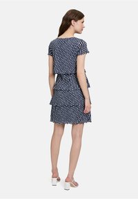 Navyblaues Kleid mit einem wellenförmigen weißen Muster, kurzen Ärmeln und gestuften Rüschen am Saum. Kombiniert mit weißen High-Heel-Sandalen.